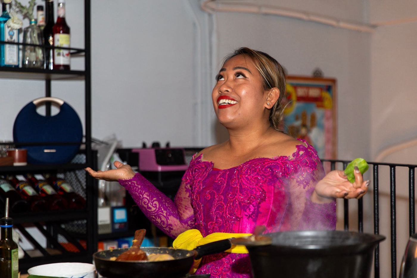 Chef Novy laughing in a magenta lace kebaya with gold sash, cooking live in a pan at a private event