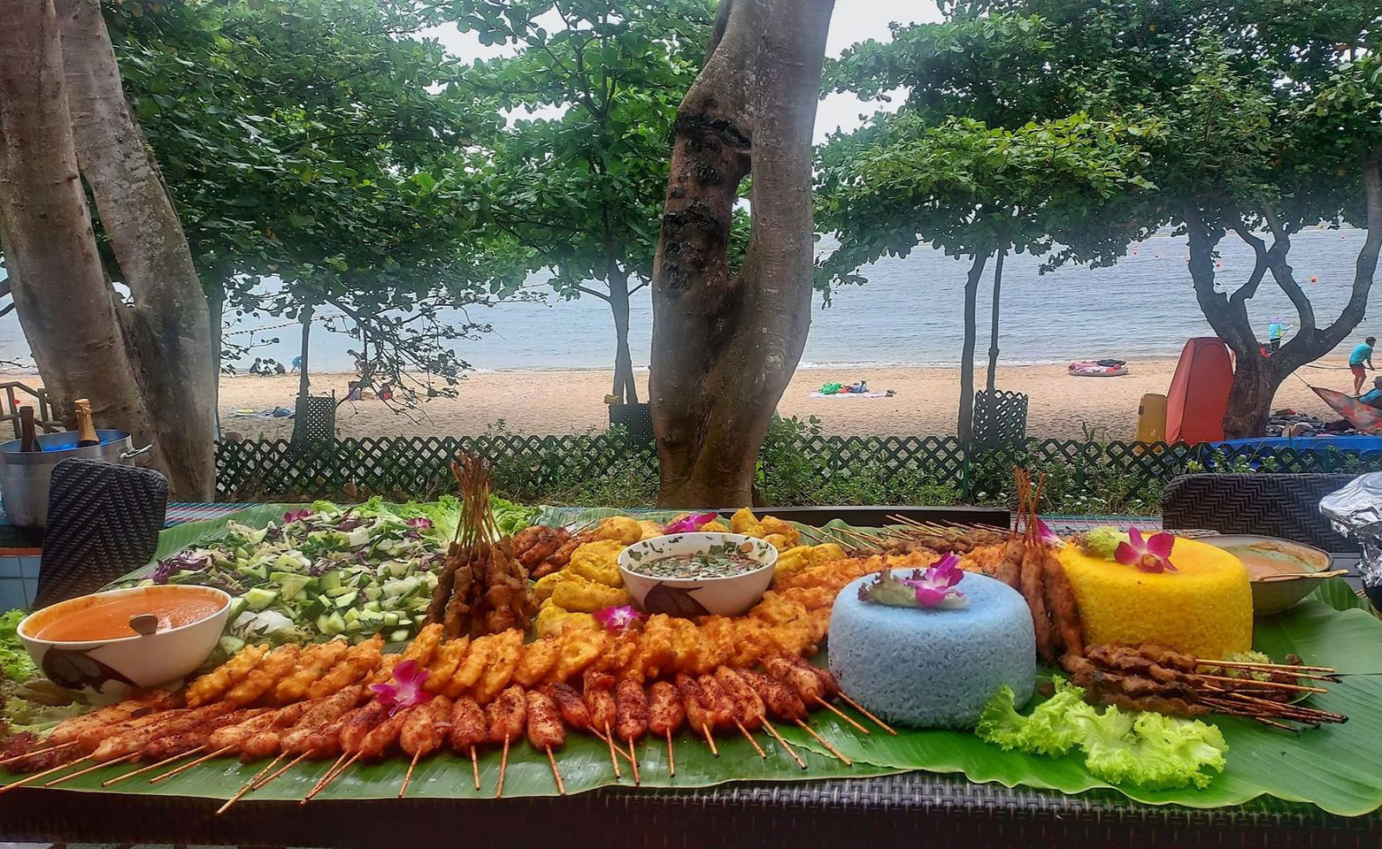 Lavish Indonesian banquet of satay skewers, golden nasi kuning, blue rice and sambals served on banana leaf with the sea and beach in the background