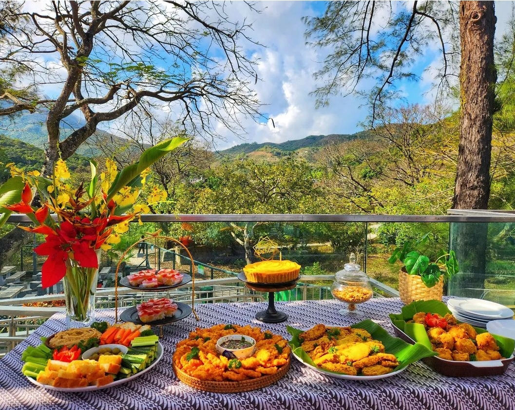Banquet table spread with golden fritters, cheesecake, satay and crudités on a batik cloth, set on a terrace with Lantau's mountains and jungle views