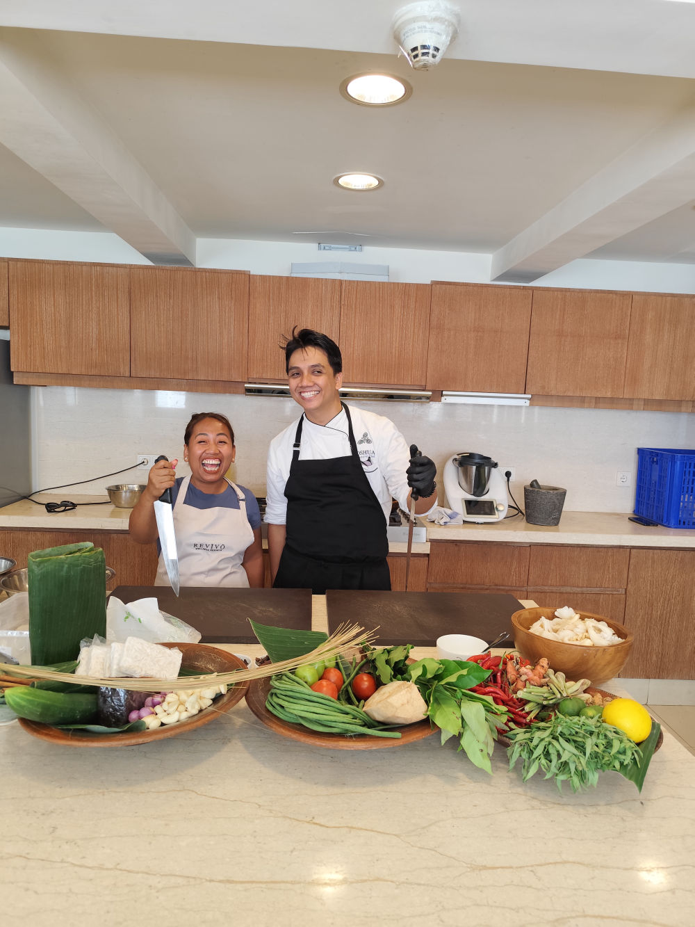 Guest Chef Novy laughing in an apron beside the resort kitchen team, with trays of fresh Indonesian produce — herbs, chilies, galangal and vegetables — laid out on the counter at Revivō Wellness Resort, Bali