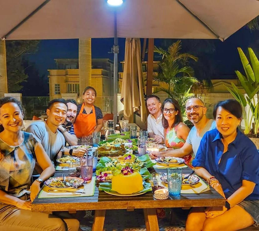 Chef Novy in an orange apron serving a full table of happy guests at a private home dinner — banana-leaf feast and nasi kuning centrepiece under a garden umbrella at night