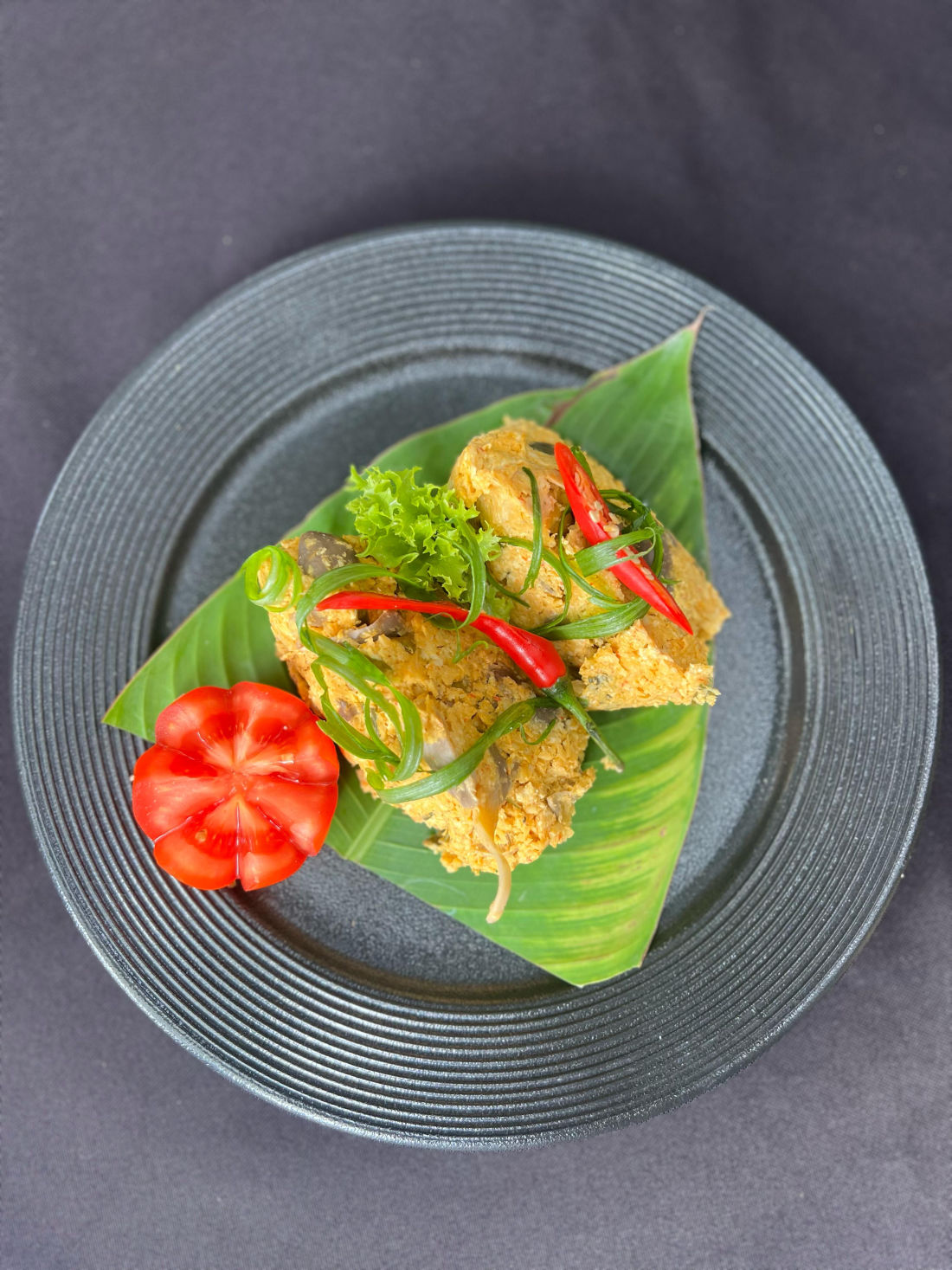 Signature plated Indonesian dish — spiced tempeh with chili on banana leaf over dark stoneware